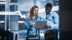 前景には2人の専門家がグラフを表示するモニターが置かれたモダンなハイテクオフィスでノートパソコン上のデータを確認しています。2人とも眼鏡をかけており、青いシャツを着ています。